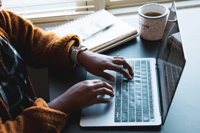 person in yellow sweater typing on laptop with notebook and mug nearby